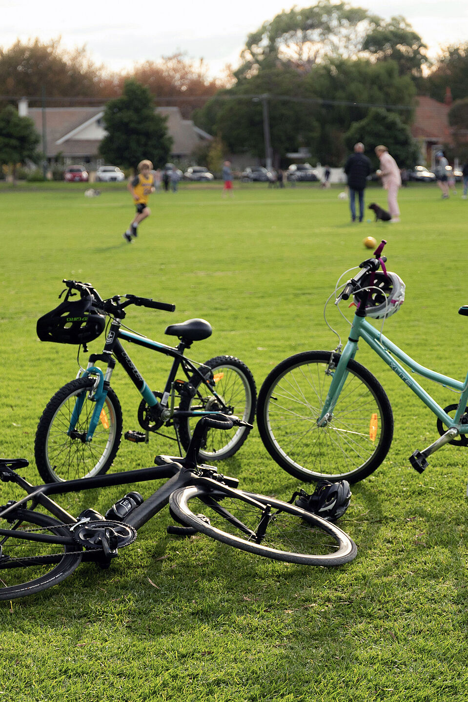Fahrräder an einem Bolzplatz Im Vordergrund sind drei Räder stehend und liegend auf einer Rasenfläche zu sehen, im Hintergrund sind ein paar Menschen auf dem Rasen stehend mit einem Fußball zu sehen.