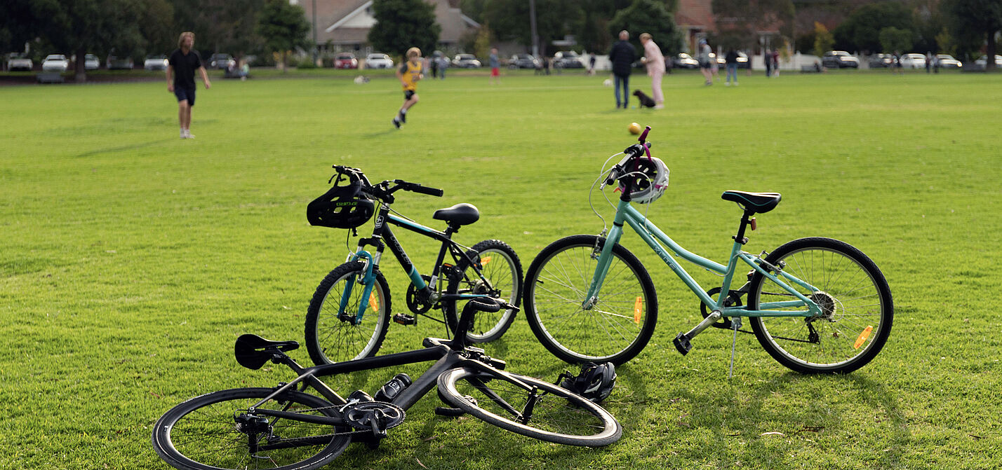 Fahrräder an einem Bolzplatz Im Vordergrund sind drei Räder stehend und liegend auf einer Rasenfläche zu sehen, im Hintergrund sind ein paar Menschen auf dem Rasen stehend mit einem Fußball zu sehen.